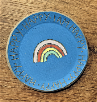 A clay plate with a light blue glaze and a rainbow design carved out. The words "I am happy" are carved into the rim of the plate.